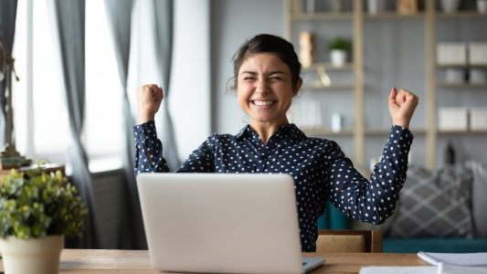 Euphoric young girl celebrates victory triumph at home desk with laptop computer